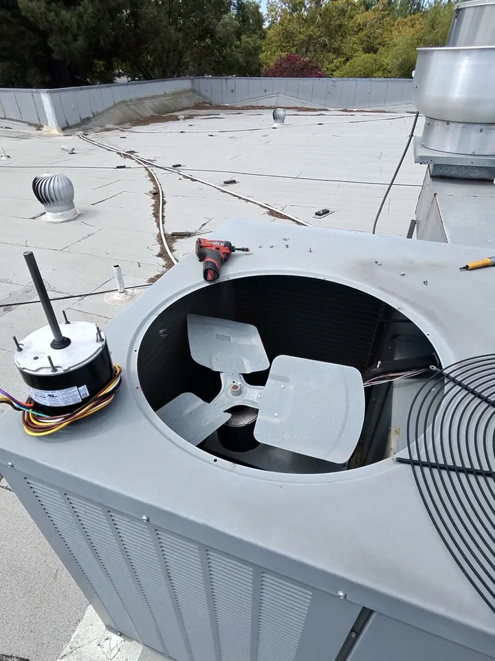 Open rooftop AC unit with the fan grille removed and a replacement condenser fan motor beside the opening